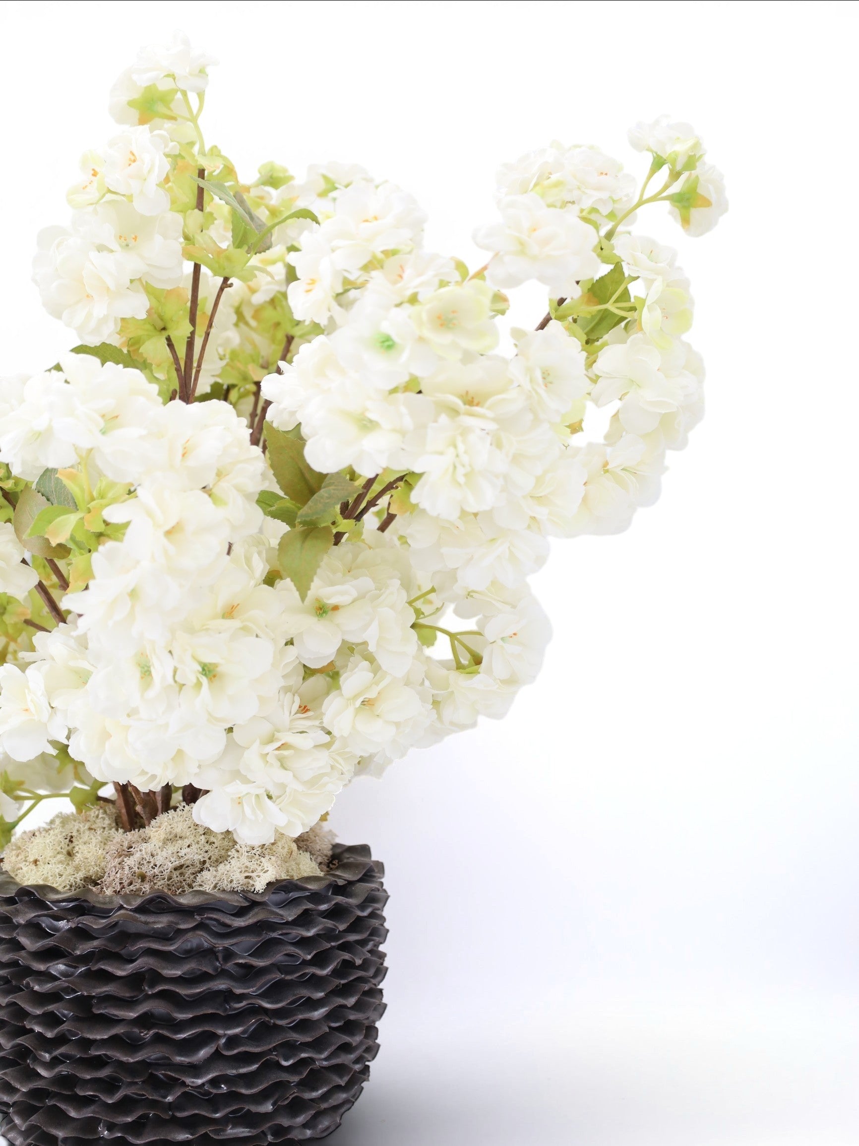 Sixty3 Styled artificial white cherry blossom arrangement in a dark textured pot with green foliage.