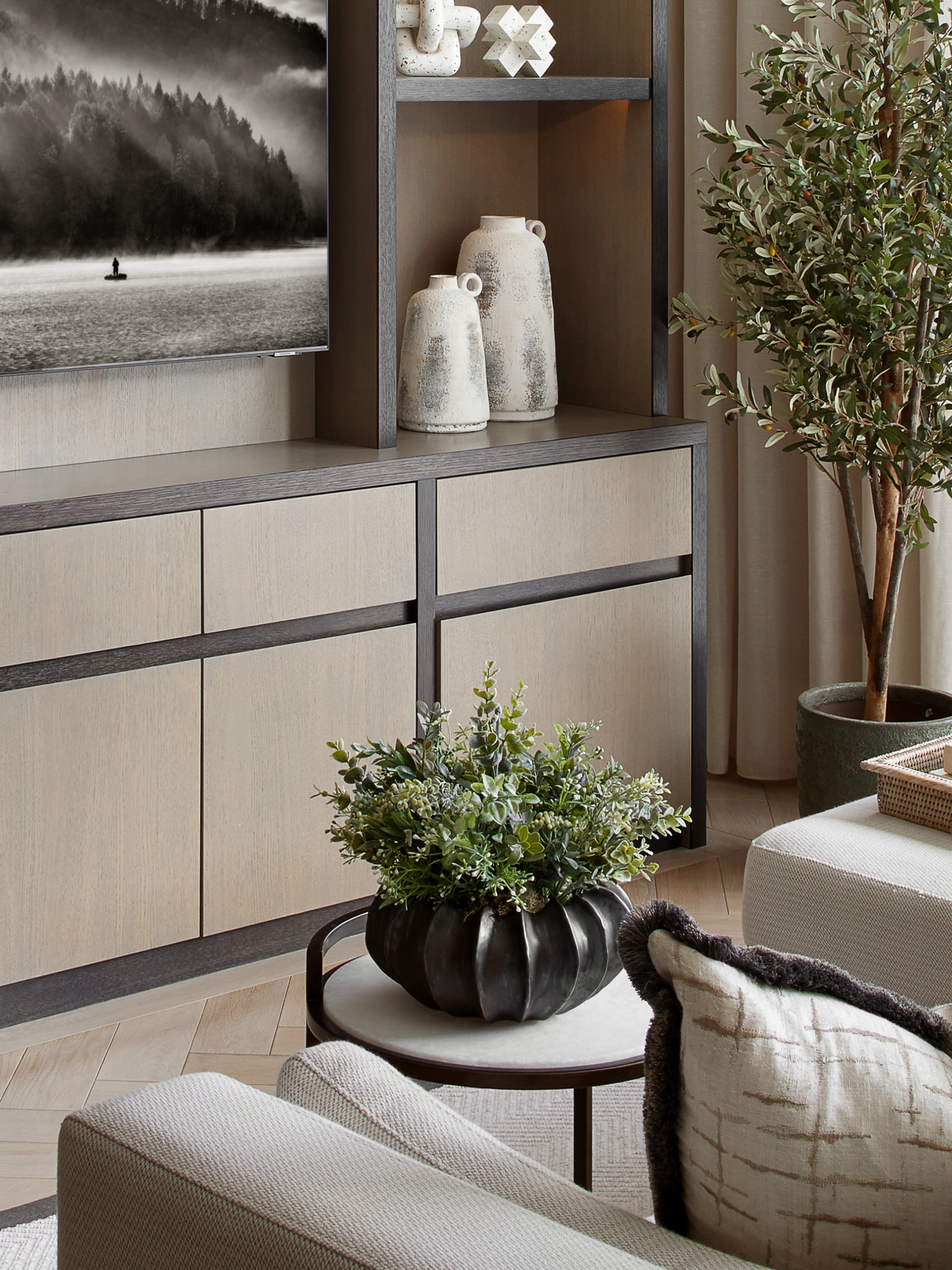 A bronze ribbed vase with a round base placed on a table, with a plant inside it, in a room with wooden cabinets and a painting in the background.